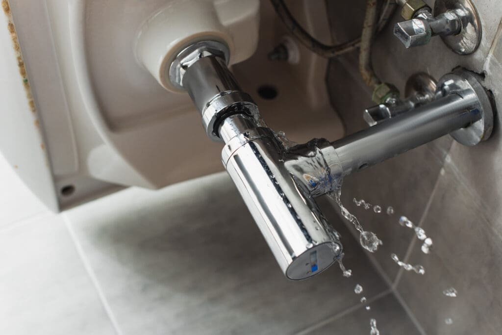 A close-up of a leaking, chrome-plated plumbing pipe under a sink showcases the need for prompt plumbing repairs. Water drips from the connector, catching the light on its shiny surface. Tiled flooring and other pipes hint at the comprehensive property services needed to prevent further issues.