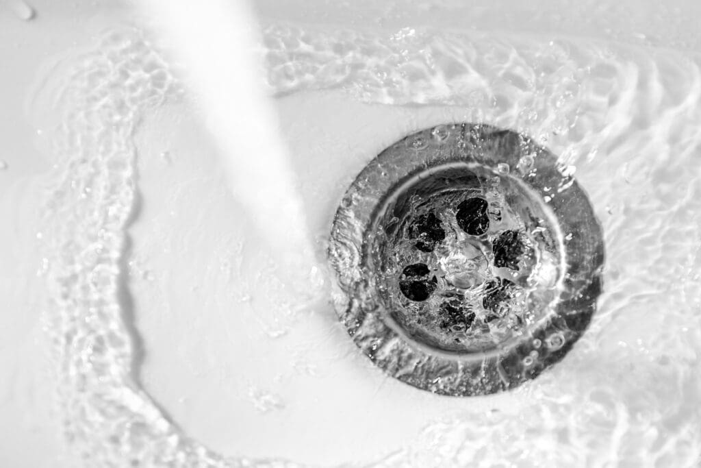 Water cascades into a stainless steel sink drain from above, perfectly captured in black and white. The image showcases the swirling motion of the water and the textured surface of the drain, evoking a sense of artistry often admired by those skilled in bathroom installations.