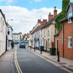 A narrow street in a quaint town with charming brick and stone buildings on both sides. A few pedestrians and a car attest to the day's calm. The sky is partly cloudy, while streetlights stand guard over green trash bins, hinting at the nearby property services bustling with shower repairs and bathroom installations.