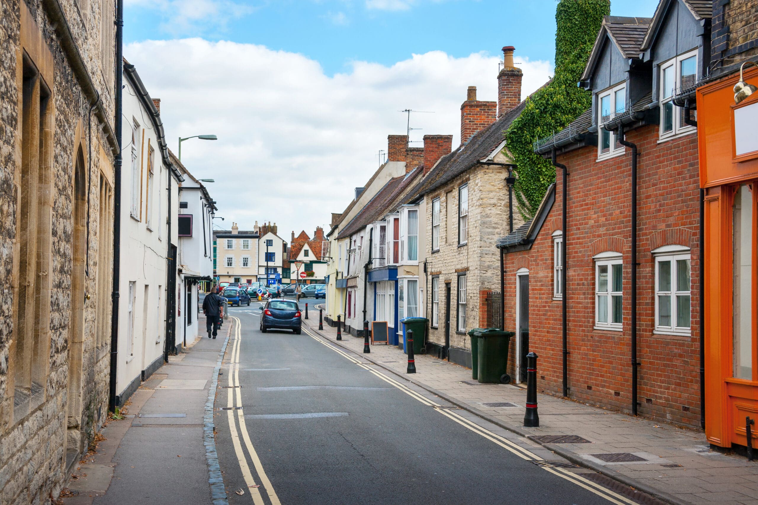 A narrow street in a quaint town with charming brick and stone buildings on both sides. A few pedestrians and a car attest to the day's calm. The sky is partly cloudy, while streetlights stand guard over green trash bins, hinting at the nearby property services bustling with shower repairs and bathroom installations.