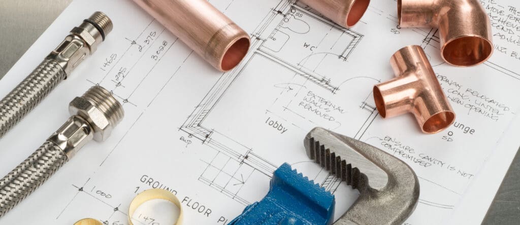 Plumbing tools and parts, essential for bathroom installations, including a wrench, copper pipes, and connectors, are laid out on architectural plans for a building's plumbing system.
