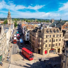 A scenic view of a bustling city street in Oxford, England, features historic architecture and several red double-decker buses. Among the people walking along the sidewalks, you might even spot a plumber heading to a nearby bathroom installation. The landscape reveals old buildings under a clear blue sky.
