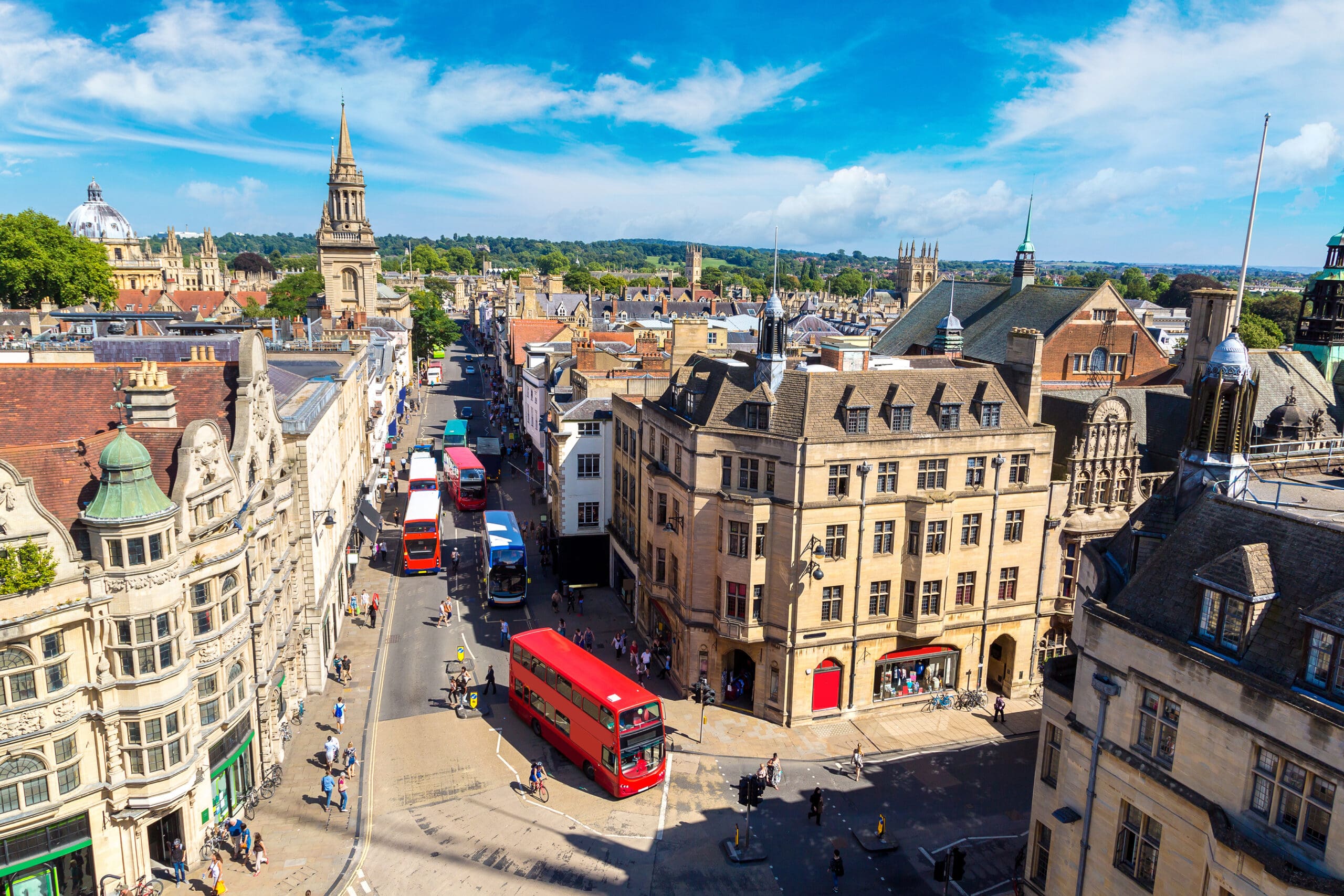 A scenic view of a bustling city street in Oxford, England, features historic architecture and several red double-decker buses. Among the people walking along the sidewalks, you might even spot a plumber heading to a nearby bathroom installation. The landscape reveals old buildings under a clear blue sky.