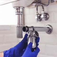 Person wearing blue gloves is expertly tackling plumbing repairs on a metal sink drain pipe under a bathroom sink. The wall, adorned with white tiles, showcases visible plumbing connections, reflecting the precision that defines quality property services.