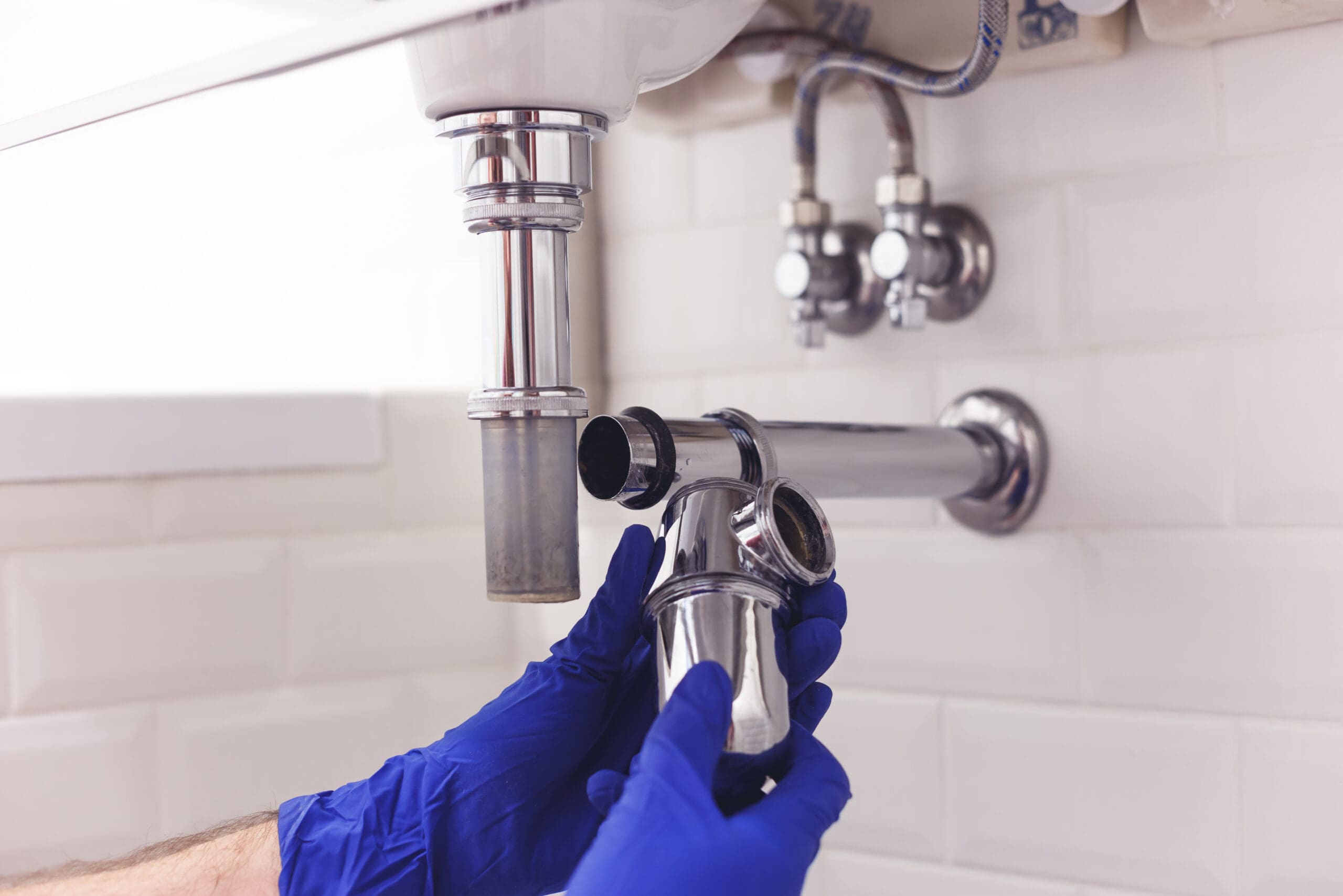 Person wearing blue gloves is expertly tackling plumbing repairs on a metal sink drain pipe under a bathroom sink. The wall, adorned with white tiles, showcases visible plumbing connections, reflecting the precision that defines quality property services.
