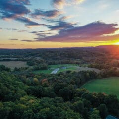 Aerial view of a sprawling landscape at sunset, with vast green fields and dense forests. The sun dips below the horizon, painting the sky in orange and purple hues. A small road leads to distant structures, perhaps undergoing plumbing repairs amid nature's serene beauty.