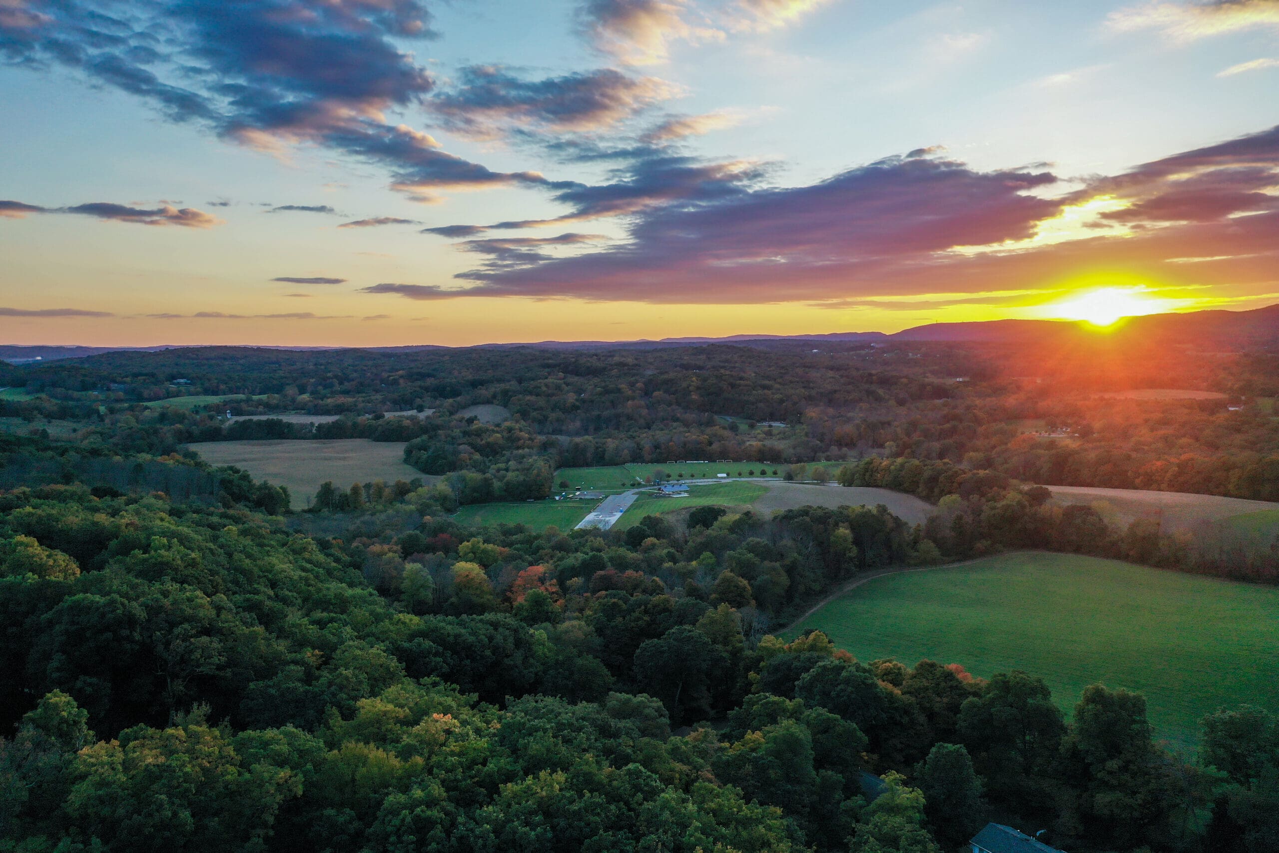 Aerial view of a sprawling landscape at sunset, with vast green fields and dense forests. The sun dips below the horizon, painting the sky in orange and purple hues. A small road leads to distant structures, perhaps undergoing plumbing repairs amid nature's serene beauty.