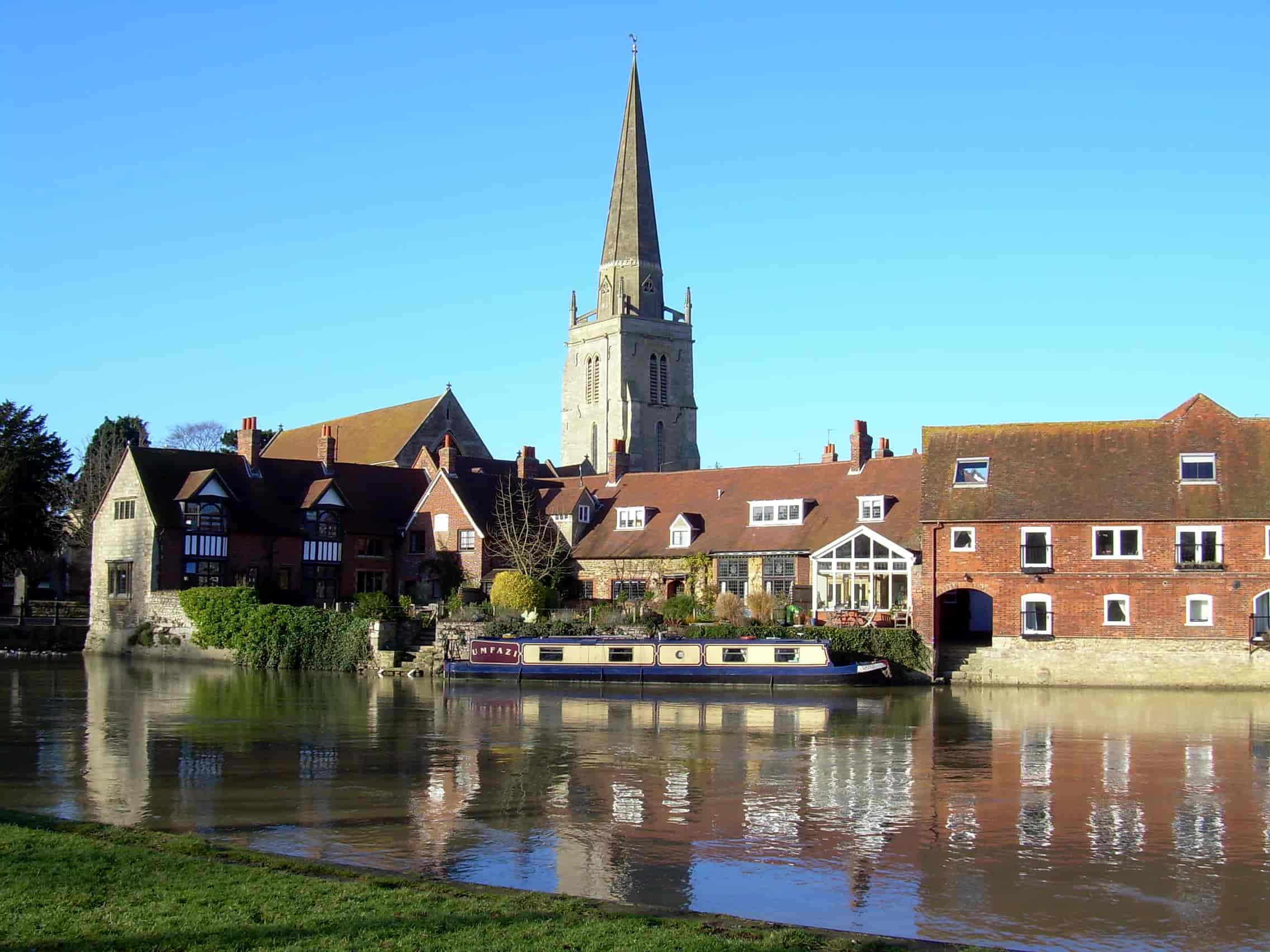 A riverside village with red-roofed buildings and a tall church steeple, reflected in calm water under a clear blue sky. A narrowboat is moored along the riverbank in front of the houses.