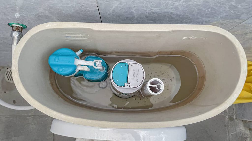 Open toilet tank showing the internal mechanisms, including the fill valve, float, and flush valve, partially submerged in water. The tank cover is removed, and some debris is visible in the water.