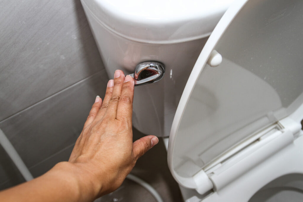 A hand is pressing the flush lever on a white toilet, with the toilet seat lid open and the flush tank visible.