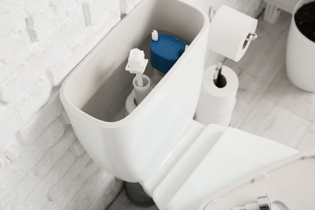 An open toilet tank showing its internal mechanisms, with the lid resting to the side. Two toilet paper rolls and a plant are nearby on a light-colored tiled floor next to a white brick wall.