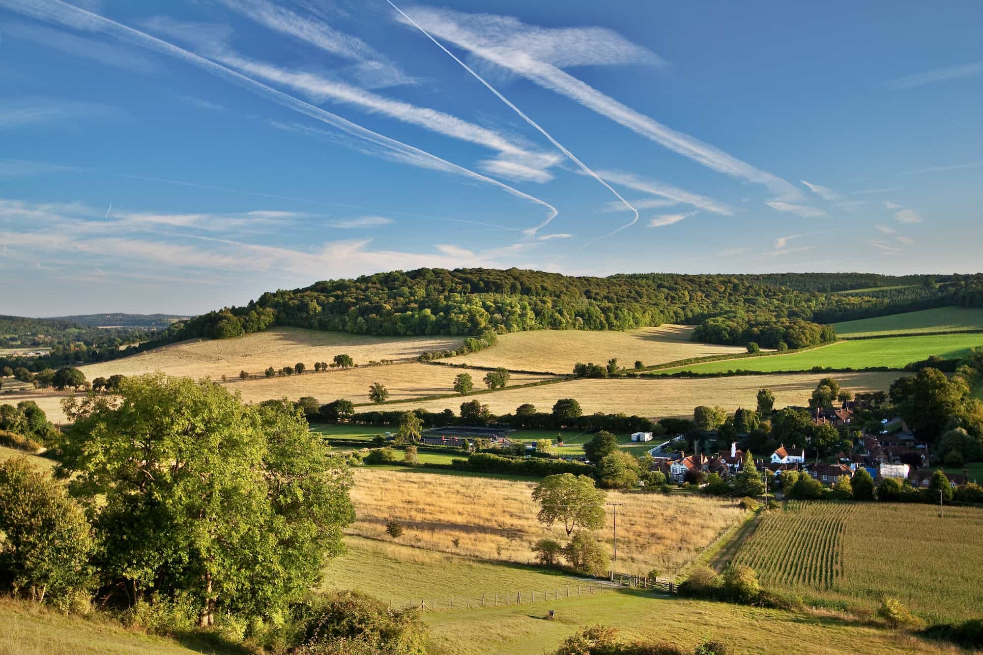 Rolling countryside with green fields, scattered trees, and a small village of houses nestled below a wooded hill. Blue sky above has several white airplane contrails crossing over the landscape.