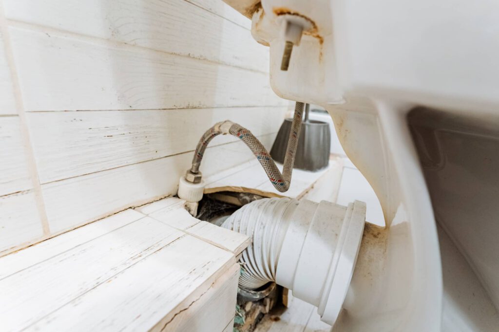 Close-up view of the back of a toilet with exposed plumbing, including a flexible water supply hose and a large white drain pipe, against a white wooden wall.