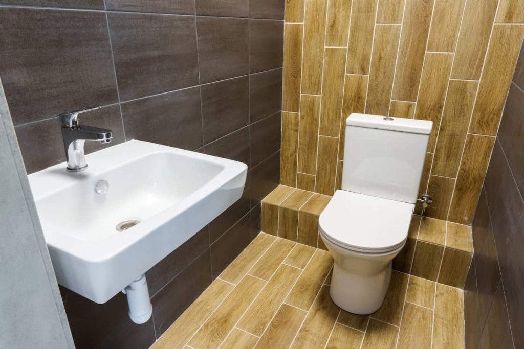 A modern bathroom with a white sink and faucet on the left and a white toilet on the right, featuring brown wood-look tiles on the floor and walls.