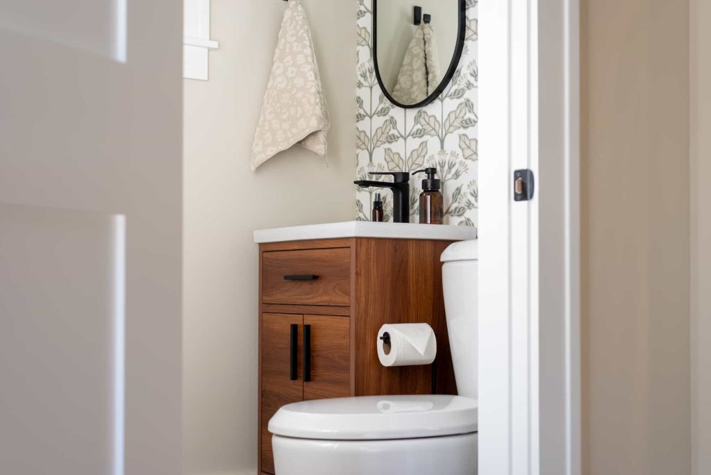 A modern bathroom with a white toilet, a wooden vanity with black handles, a round mirror, patterned wallpaper, a towel hanging on the wall, liquid soap dispensers, and a roll of toilet paper on a holder.