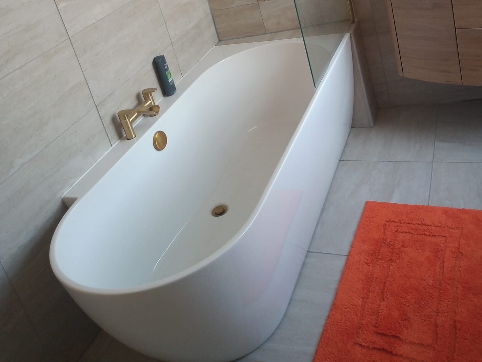 A modern bathroom featuring a white freestanding bathtub with gold fixtures and a glass shower partition. The space includes a wooden vanity with a built-in sink, a mirror above, and an orange bath mat. Light beige tiles adorn the walls, completing this exquisite example of premium bathroom installations.